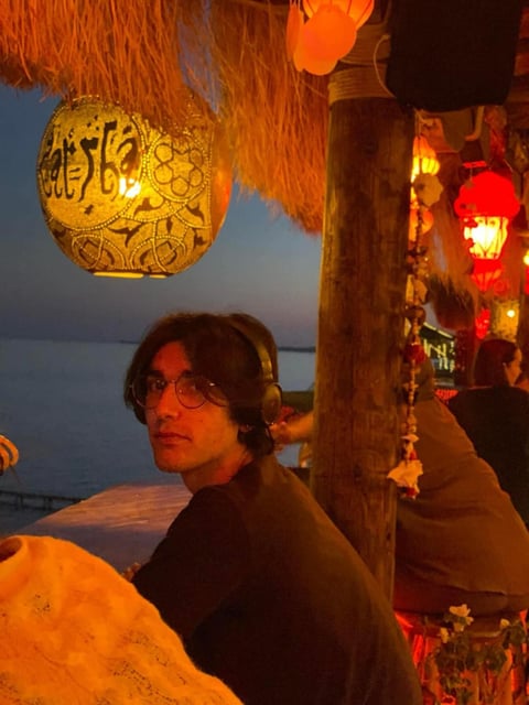 Man wearing glasses sitting at waterfront at dusk, surrounded by glowing decorative lanterns and wooden posts