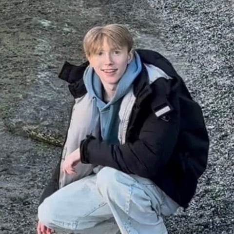 Young person with blonde hair wearing a black jacket, blue hoodie, and light jeans, sitting against a concrete wall